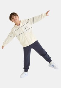 Felpa con cappuccio beige chiaro con zip, strisce nere e logo, abbinata a pantaloni da jogging blu navy scuro e sneaker bianche con dettagli blu.