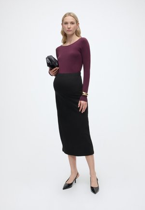 Frau in einem enganliegenden burgunderroten Langarmshirt, schwarzem Midirock, schwarzen High Heels, hält eine schwarze Clutch und steht vor einem schlichten weißen Hintergrund.