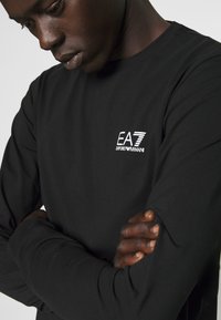 Maglietta nera a maniche lunghe realizzata in tessuto morbido, con logo EA7 bianco sul petto. Design aderente con una texture liscia e scollo rotondo.