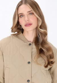 En blond kvinde med bølget hår iført en beige quiltet jakke med metalknapper mod en ensfarvet hvid baggrund.