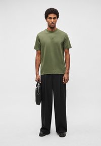 Olivzaļš īsroku T-krekls ar apaļu kakla izgriezumu, kopā ar vaļīgām melnām biksēm. Modelis tur melnu somu ar sudraba sprādzi.