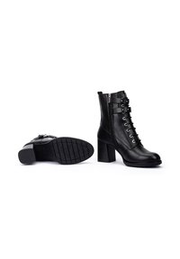 Botas negras de cuero hasta el tobillo con tacón cuadrado, parte frontal con cordones y cremallera lateral. Presenta ojales de metal y una suela de goma texturizada.