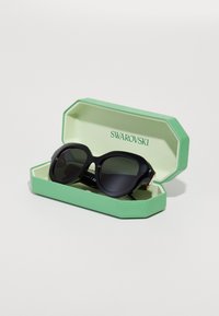 Ochelari de soare negri cu o ramă groasă, depozitați într-o cutie octogonală verde. Interiorul este verde deschis, cu "SWAROVSKI" imprimat în verde închis.