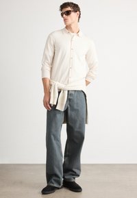 Beige gombos kardigán hosszú ujjakkal egy póló felett, laza kék farmernadrággal és fekete cipővel párosítva. A modellen napszemüveg van.
