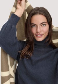 Marinefarbener Rollkragenpullover aus strukturiertem Material; das Model hält einen beigen und cremefarbenen Schal mit geometrischem Muster und sanftem Fall.