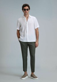 Chemise blanche en lin à manches courtes, pantalon olive, et chaussures gris clair à enfiler. Individu portant des lunettes de soleil, debout contre un fond neutre.