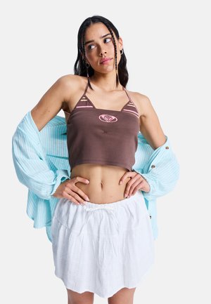 Jeune femme avec les cheveux tressés porte un crop top marron, une jupe blanche et une chemise à rayures bleu clair drapée sur les épaules, posant les mains sur les hanches.