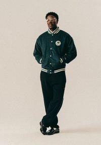 Mees, kes kannab musta ülikooli jakki embleemiga, musti pükse, loafer-susse ja prille, seisab jalad risti seina vastu lihtsas taustas.