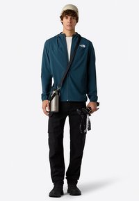 Veste zippée turquoise avec capuche, associée à un pantalon cargo noir. Accessoires comprenant une bouteille d'eau grise et des objets noirs à la main. Chaussures décontractées.