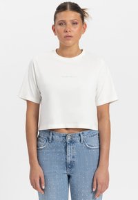 Korte, cropped witte t-shirt met korte mouwen, voorzien van een kleine multikleuren tekstdesign op de voorkant. In combinatie met lichtblauwe jeans met versieringen.