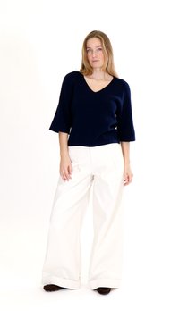 Kvinde stående iført en marineblå V-hals top med albuelange ærmer, brede hvide bukser og mørke sko mod en ensfarvet hvid baggrund.