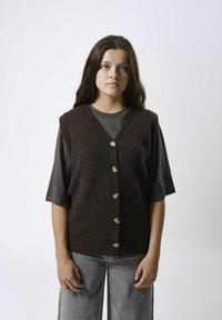 Dunkelbrauner gestrickter ärmelloser Pullover mit V-Ausschnitt, der sechs goldene Knöpfe auf der Vorderseite und einen gerippten Saum aufweist. Trägt man über einem dunkelgrauen Hemd.
