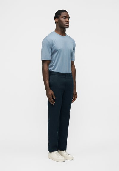 Άνδρας που στέκεται φορώντας ανοιχτό μπλε κοντομάνικο πουκάμισο, σκούρο μπλε παντελόνι και λευκά αθλητικά παπούτσια μπροστά σε απλό γκρι φόντο.