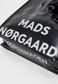 Fekete, fényes táska, nagy fehér felirattal: "MADS NØRGAAARD COPENHAGEN." Simább anyagú, téglalap alakú és enyhén fénylő.
