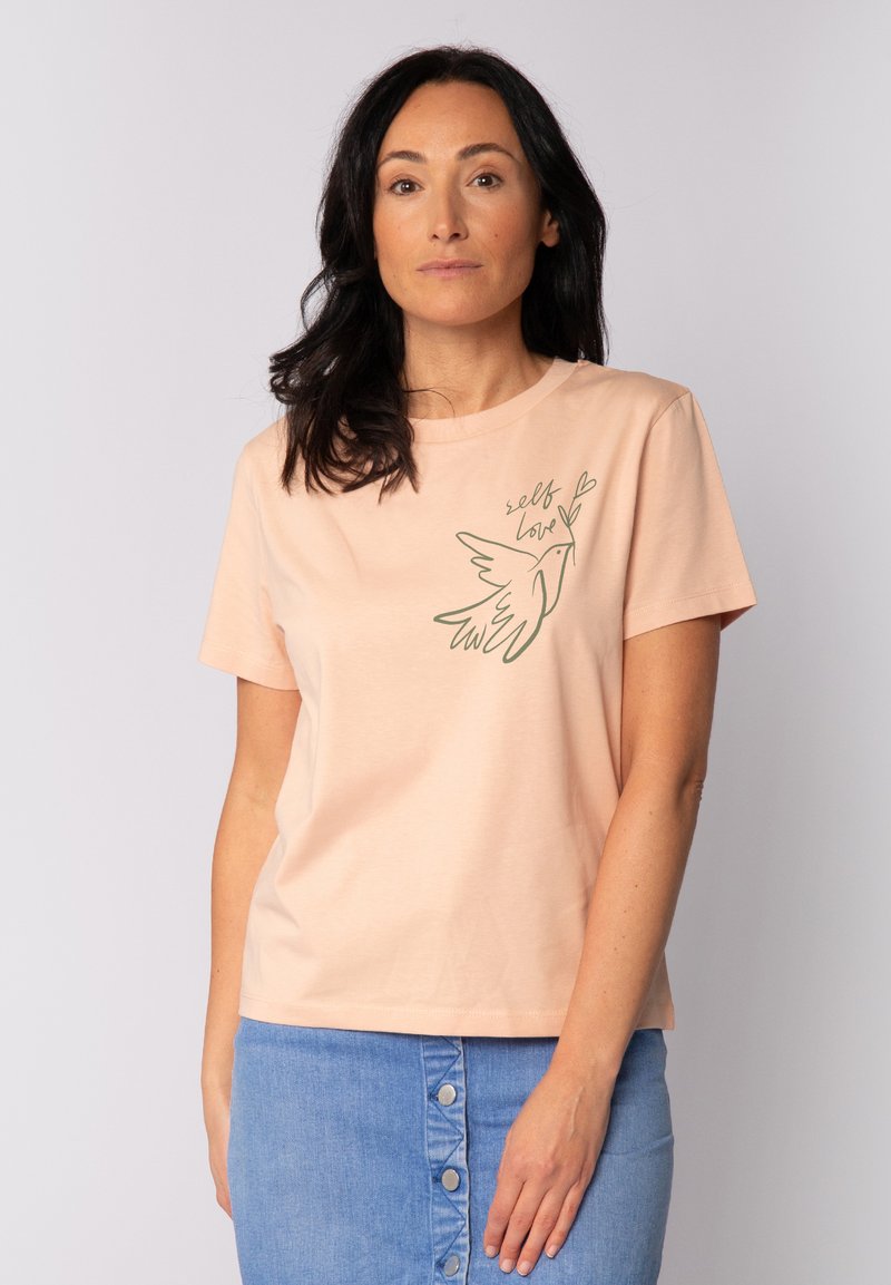 T-shirt en coton couleur pêche avec des manches courtes et une coupe décontractée, comportant un graphique vert d'un oiseau et le texte "self love" sur la poitrine.