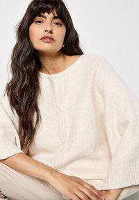 Světle béžový pletený svetr s širokými rukávy, který má jemnou strukturu a středový šev. Stylizovaný s neutrálně tónovanými kalhotami.