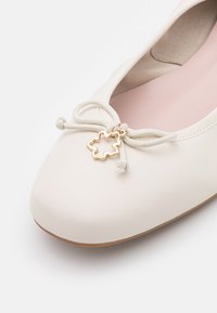 Chaussure plate de ballet blanche avec un nœud et un pendentif doré en forme de feuille sur le dessus, à bout arrondi et doublure intérieure beige clair.