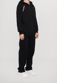 Černá mikina s kapucí s logem a pruhovanými detaily na kapuci. Kombinováno s černými kalhotami a bílými teniskami. Hladká textura látky.