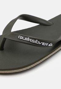 Fekete flip-flop, texturált talppal, vékony, bordázott dizájnnal rendelkező pántokkal és fehér logóval. Barna talppal, fehér cérnával díszítve.