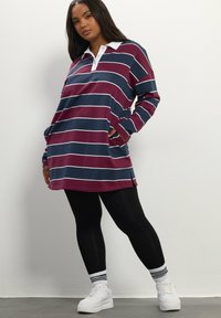 Übergroßes Rugby-Shirt im maroonfarbenen und navy gestreiften Design, mit einem weißen Kragen, langen Ärmeln und seitlichen Schlitzen. Kombiniert mit schwarzen Leggings und weißen Turnschuhen.