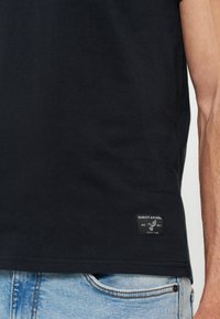 T-shirt noir avec une petite étiquette "Quality Apparel" près de l'ourlet, porté avec un jean bleu clair, vue partielle du torse et du bras d'une personne.