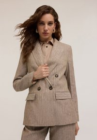 Blazer a rayas beige de doble botonadura con dos bolsillos frontales y botones marrones, combinado con una camisa de color beige claro con cuello.
