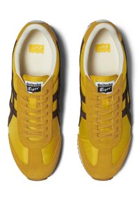 Žluté semišové a nylonové tenisky s hnědými pruhy, texturovanými tkaničkami a polstrovaným límcem. Branding viditelný na jazyku. Klasický sportovní design.