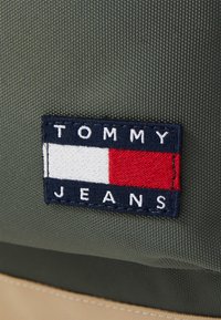 Žalia audinio krepšys su tamsiai mėlyna siuvinėta etikete "TOMMY JEANS", baltomis ir raudonomis horizontaliomis juostomis bei tekstūruotu fonu.