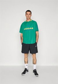 Πράσινο t-shirt Adidas με κίτρινο λογότυπο, μαύρο σορτς, λευκές κάλτσες και μαύρα αθλητικά παπούτσια. Διαθέτει κοντά μανίκια και άνετη εφαρμογή.