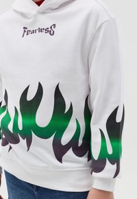 Witte hoodie met "Fearless" in paarse tekst, versierd met groene en zwarte vlamgraphics aan de onderrand. Het materiaal lijkt zacht en heeft een relaxte pasvorm.