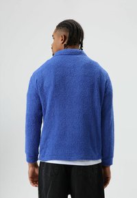 Blauwe fleece trui met een gestructureerd, zacht oppervlak en een kraag. Voorzien van lange mouwen en een losse pasvorm, gecombineerd met een donkere broek.