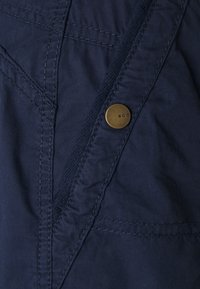 Tela azul marino con una textura suave presenta costuras diagonales y un botón redondo de latón con letras grabadas.