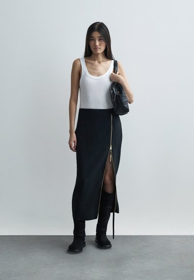 Kobieta stoi w pomieszczeniu, ubrana w białą bezrękawnik, czarną spódnicę z rozcięciem z przodu, czarne botki i trzyma czarną torbę na ramieniu.