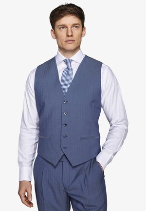 Mężczyzna ubrany w niebieską kamizelkę i spodnie garniturowe, pod którymi ma białą koszulę z niebieskim wzorzystym krawatem, stoi z jedną ręką w kieszeni.