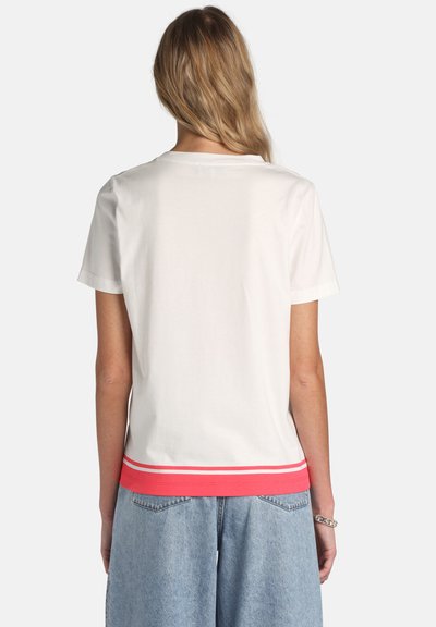 Woman with long blonde hair wearing a white short-sleeve shirt with red and white striped hem and light blue denim pants, shown from the back.