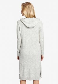 Femme blonde portant une robe sweat à capuche gris clair, se tenant de dos avec les bras détendus le long du corps.