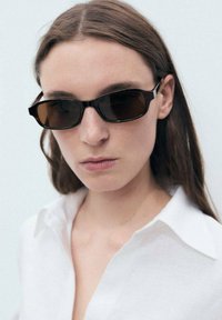 Sonnenbrille mit rechteckiger Form, dunkelbraunen getönten Gläsern und dünnen schwarzen Rahmen. Das Modell trägt ein weißes Hemd mit Kragen und hat die Haare herunter.