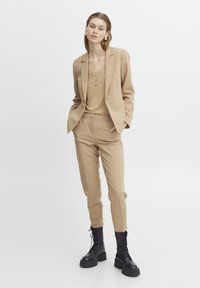 Beige op maat gemaakt pak met slim-fit broeken en een bijpassende blazer, gelaagd over een aansluitende tan top met kantdetails; zwarte chunky laarzen.