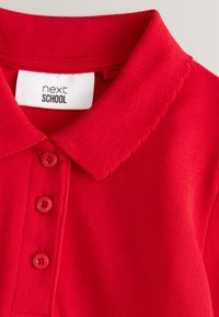 Crvena polo majica s rubom ovratnika u obliku valova, tri usklađena gumba sprijeda i bijela oznaka "next SCHOOL" unutar izreza ovratnika.