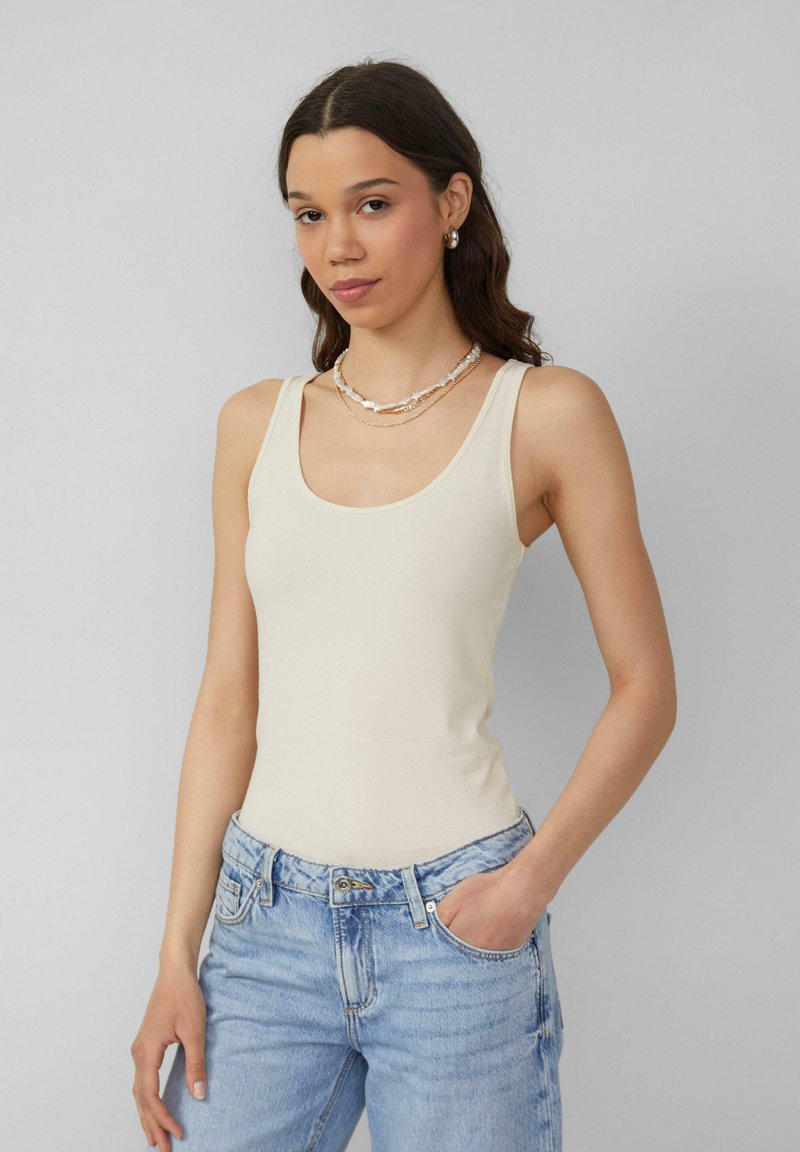 Beige getailleerde tanktop met brede banden, een gladde textuur en een ronde hals, gecombineerd met lichtblauwe jeans.