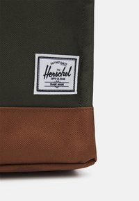 Ciemnozielona torba z brązowym spodem. Ma prostokątny kształt i białą metkę z czarnym tekstem oraz dekoracyjną ramką.