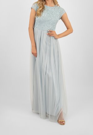 Vrouw draagt een lichtblauwe jurk met korte mouwen, een lijfje met pailletten en een lange geplooide tule rok, gecombineerd met gouden sandaal met open tenen.