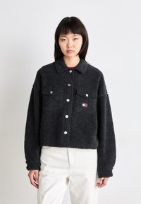 Jachetă din fleece neagră cu guler, două buzunare frontale, nasturi metalici argintii și un mic patch cu logo. Design scurt și textură moale.