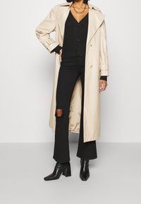 Beżowy długi płaszcz z dużymi klapami i złotymi guzikami na czarnym dzianinowym kardiganie. Czarne rozszerzane spodnie z podartymi kolanami i czarne botki na obcasie.