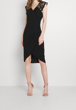 Kobieta w czarnej sukience kopertowej z koronkowymi krótkimi rękawami i cielistych szpilkach z paskiem przy kostce na jednolitym tle.