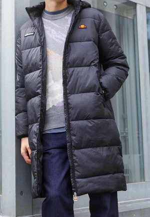 Person iført en lang sort dunjakke over en gråmønstret sweater og mørkeblå bukser, stående udendørs nær glas og beton.