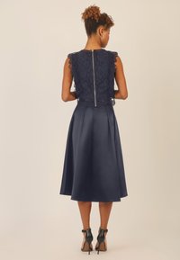 Abito blu navy con parte superiore in pizzo con bordi ondulati, corpino aderente e gonna a ruota in satin. Chiusura posteriore con zip dettagliata.