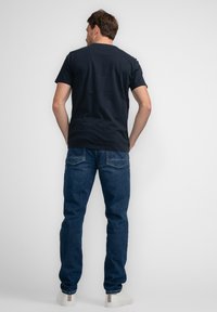 Marineblauwe katoen T-shirt met korte mouwen, gecombineerd met blauwe denim jeans en witte sneakers, van achter gezien tegen een effen achtergrond.