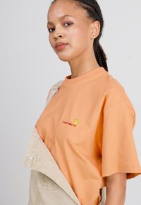 T-shirt en coton à manches courtes, de couleur pêche, avec un petit logo jaune, superposé sous une salopette beige avec une poche sur le devant.