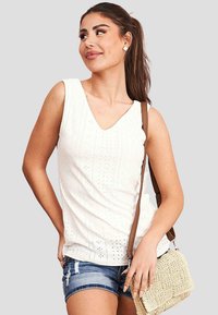 Top bianco senza maniche con scollatura a V, caratterizzato da un motivo testurizzato e piccoli fori. Abbinato a pantaloni corti in denim blu e una piccola borsa crema.